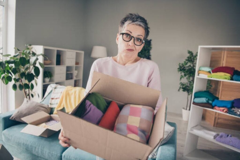Older woman with short gray hair and glasses holding a cardboard box of household items while decluttering a room.