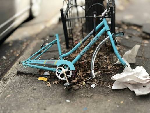 Abandoned turquoise bicycle frame missing wheels and parts, lying among leaves and trash beside a sidewalk.