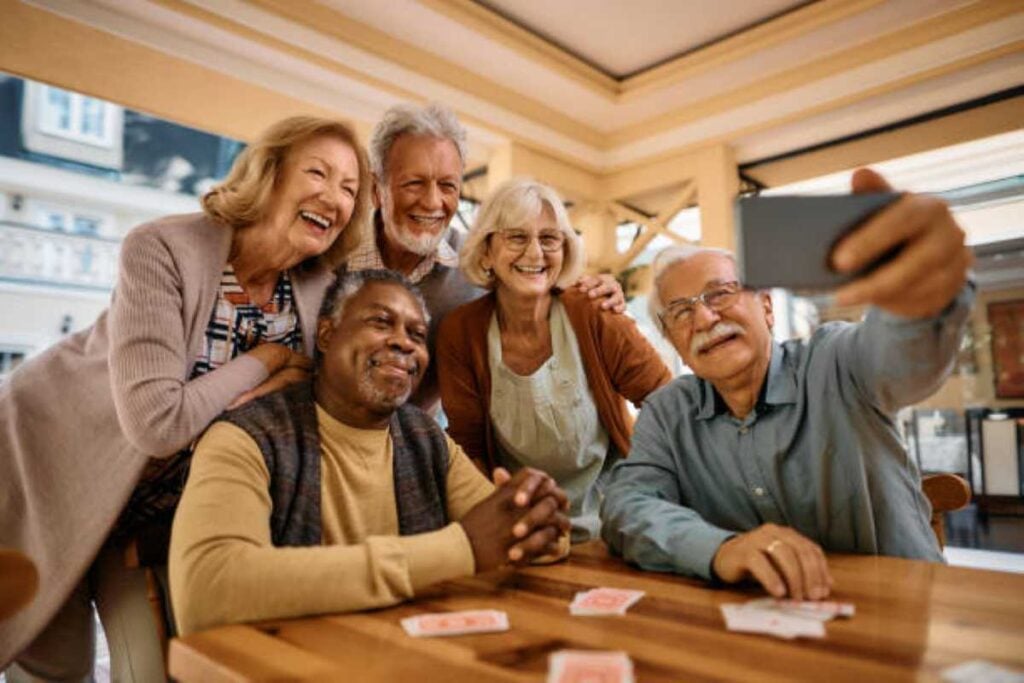 Five smiling older adults gathered around a table, laughing and posing for a group selfie while playing cards together.