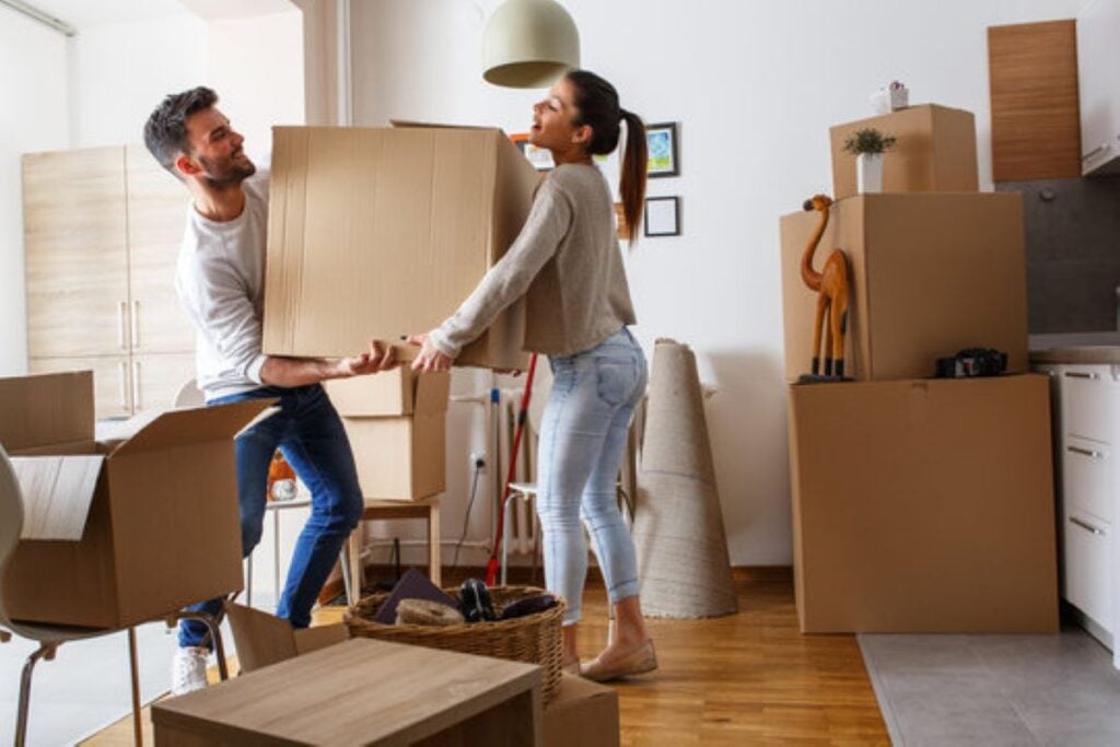 A couple carries a large cardboard box together in a bright apartment filled with stacked moving boxes and household items.