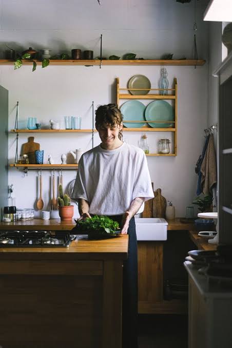 A man prepares fresh leafy greens in a cozy kitchen, surrounded by open shelves, dishes, and cooking utensils.