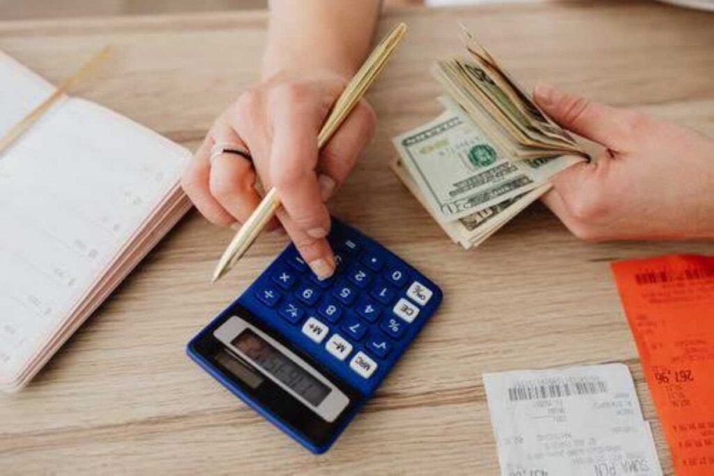 A person counts cash while using a calculator beside receipts and a notebook to track expenses.