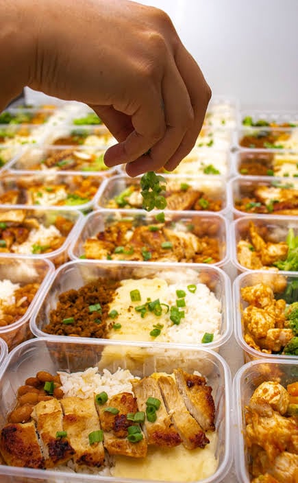 A hand sprinkles fresh herbs over rows of prepared meal containers filled with rice, chicken, vegetables, and other cooked dishes.