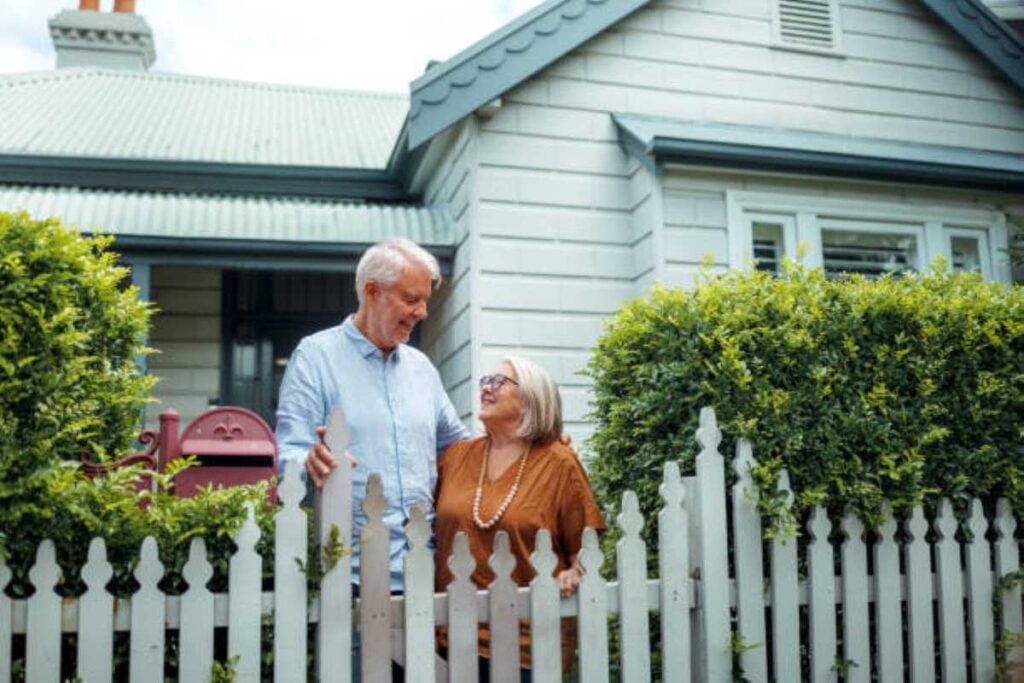 An older couple smiles at each other while standing behind a white picket fence in front of a charming traditional home.