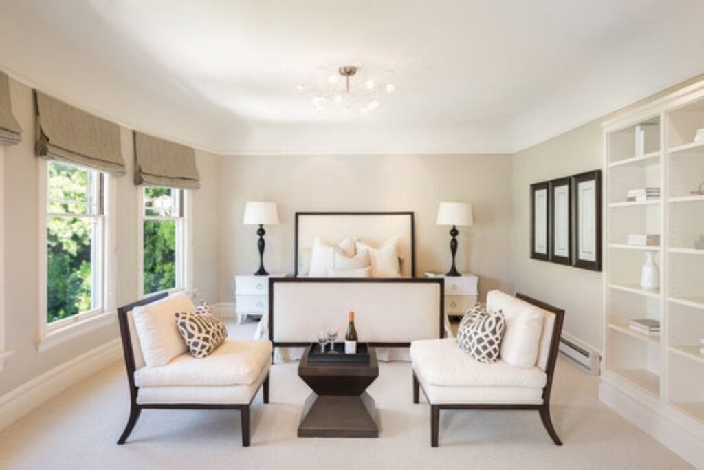 Bright neutral bedroom with two upholstered chairs, a black coffee table, large windows, built-in shelving, and a cream bed with matching bedside lamps.