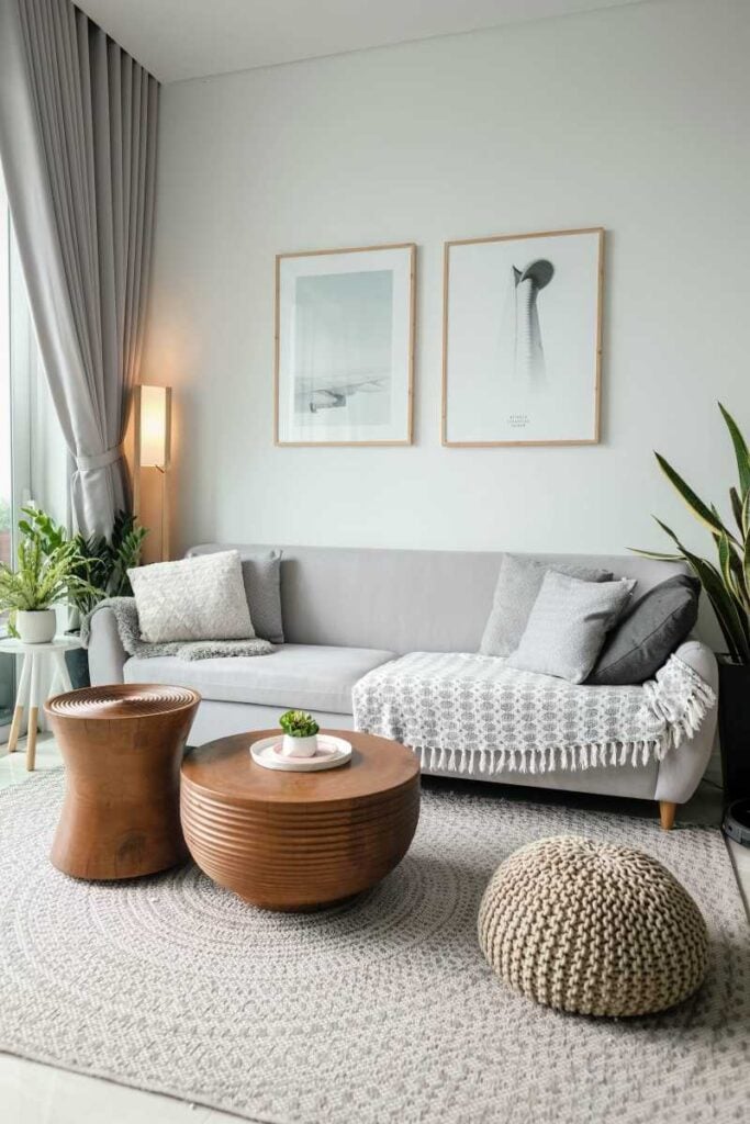 Cozy modern living room with a gray sofa, wooden coffee tables, neutral rug, framed wall art, floor-length curtains, and indoor plants.