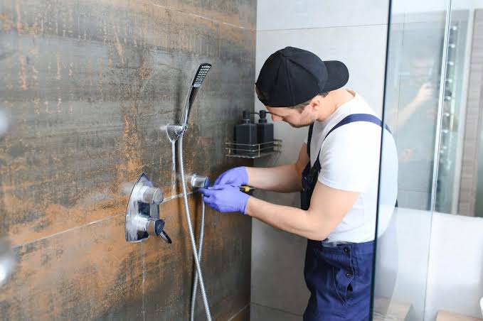 Plumber repairing a wall-mounted shower fixture in a modern tiled bathroom while wearing gloves and work overalls.