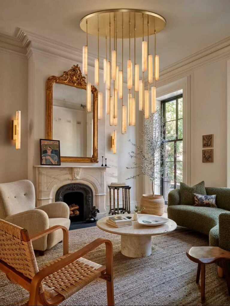 Elegant living room with a marble fireplace, ornate gold mirror, sculptural hanging chandelier, green sofa, woven chair, and round stone coffee table.