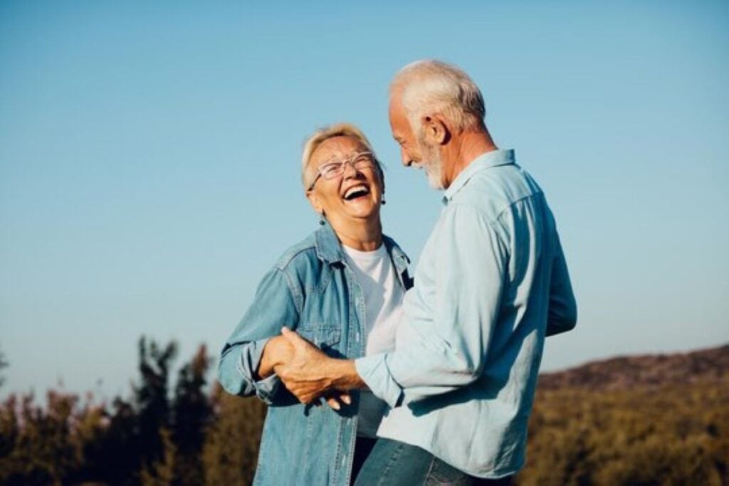 Smiling older couple holding hands and laughing together outdoors in a sunny countryside setting.