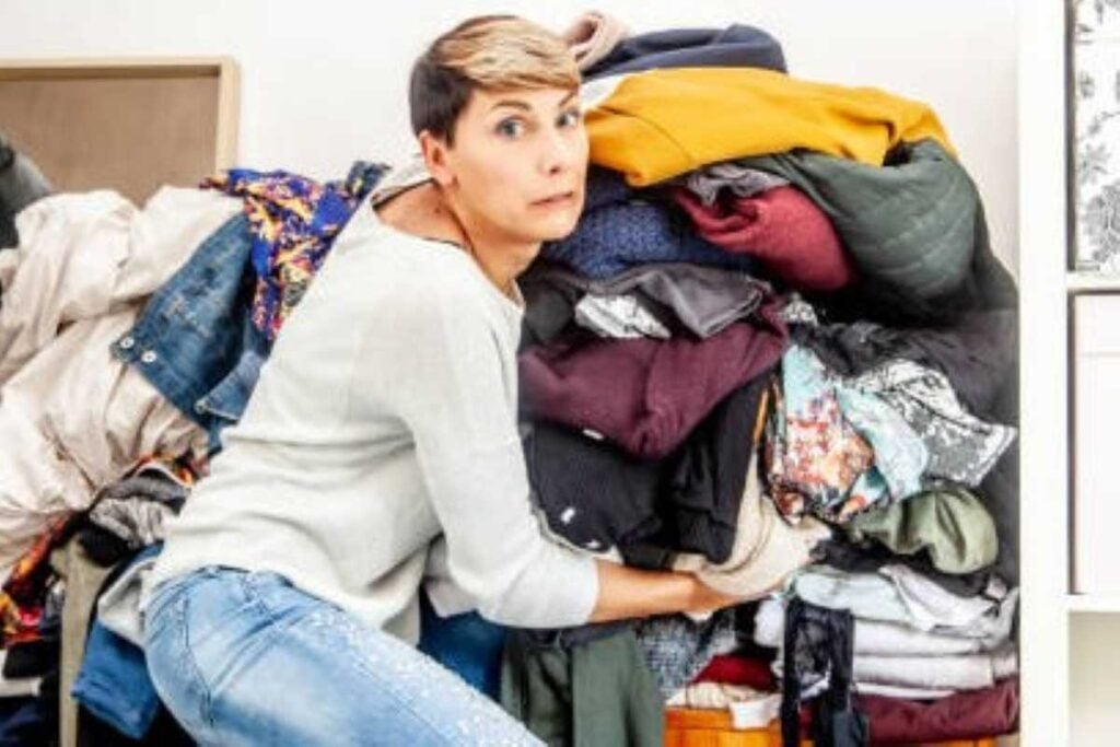A woman looks overwhelmed while trying to manage a large, overflowing pile of clothes stuffed into a closet.