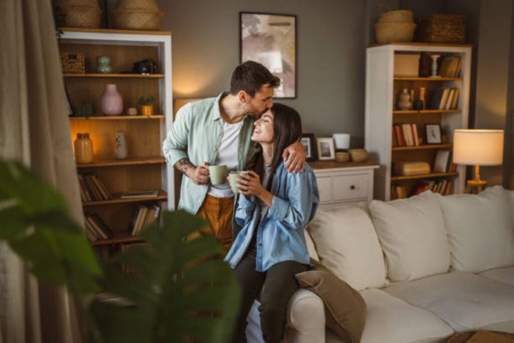 Couple sharing a warm moment while holding coffee mugs in a cozy living room with a white sofa, bookshelves, soft lighting, and houseplants.