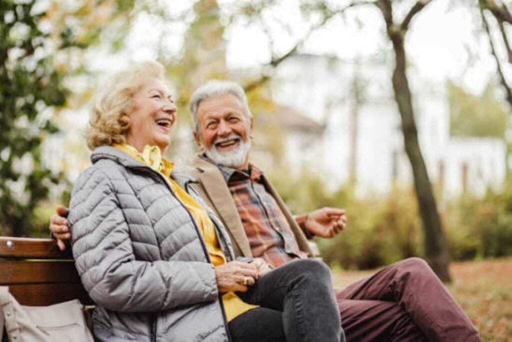 An elderly couple sitting on a park bench, laughing together and enjoying a cheerful moment outdoors surrounded by trees.