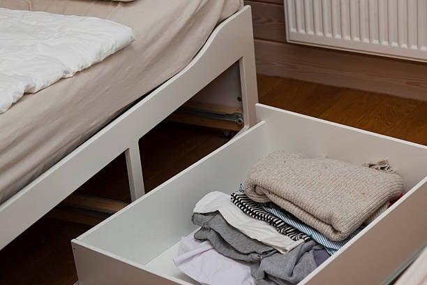 Open under-bed storage drawer filled with neatly folded clothes beneath a white bed.