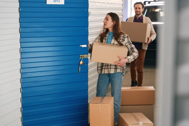 Couple carrying cardboard boxes into a storage unit with more boxes stacked nearby.