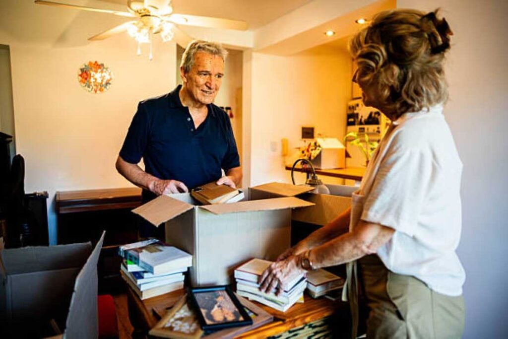 An older man and woman pack books into cardboard boxes on a table inside a warmly lit home, appearing to sort and declutter their belongings.