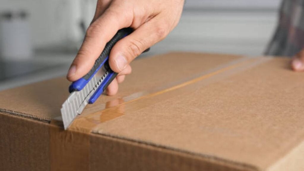 Person using a box cutter to slice packing tape on a cardboard moving box.