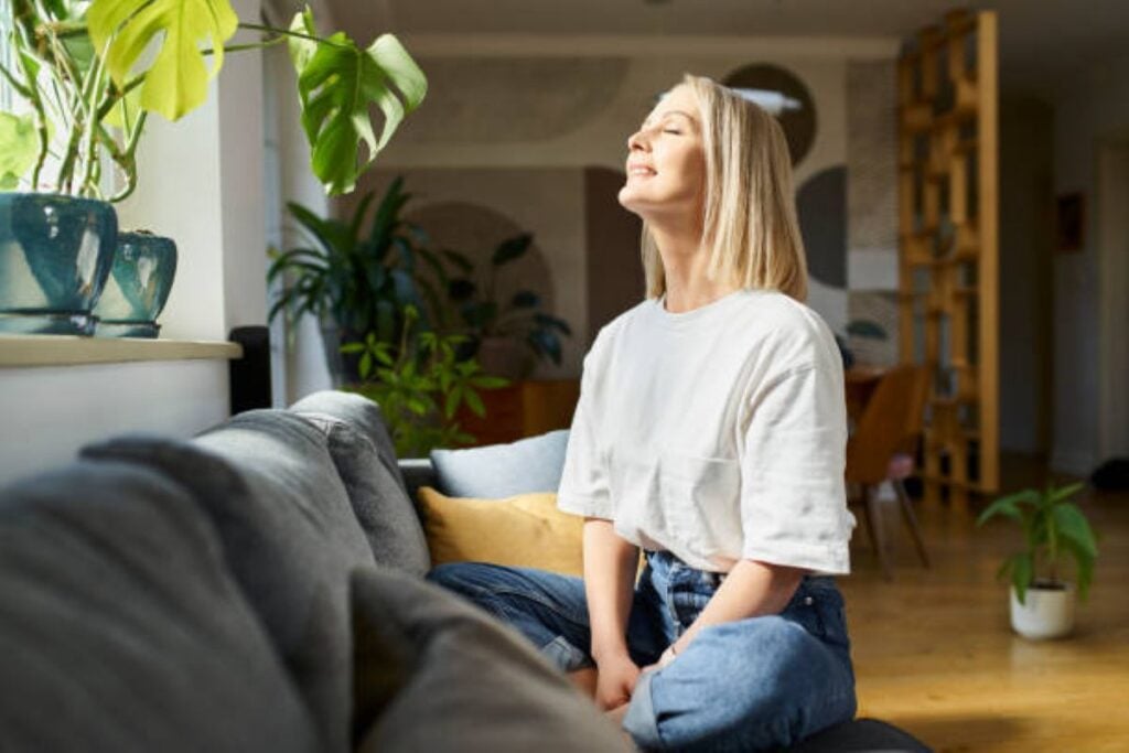 A woman sitting peacefully beside a sunlit window in a cozy, plant-filled living room, tilting her face toward the natural light.