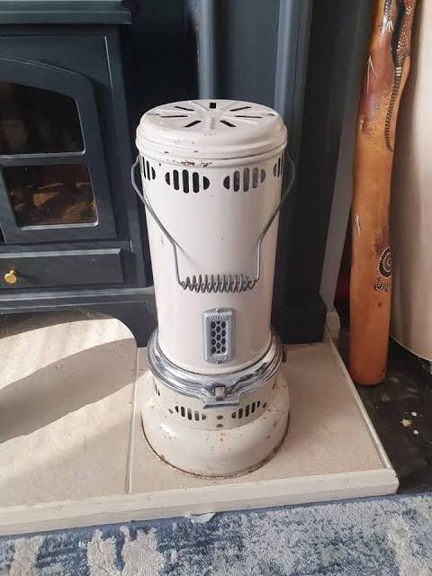 Vintage white cylindrical paraffin heater with a wire handle, standing beside a black wood-burning stove on a tiled hearth.