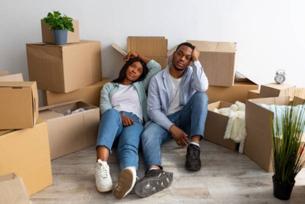 Two tired people sitting on the floor surrounded by cardboard moving boxes in a nearly empty room.