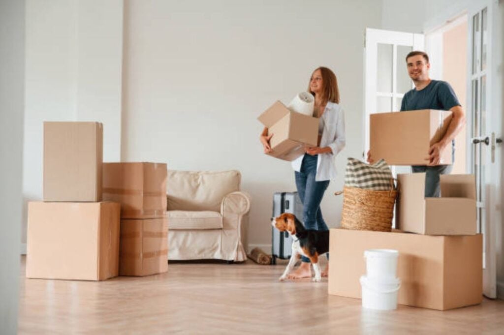 Couple carrying moving boxes into their new home with their dog, surrounded by packed cardboard boxes.