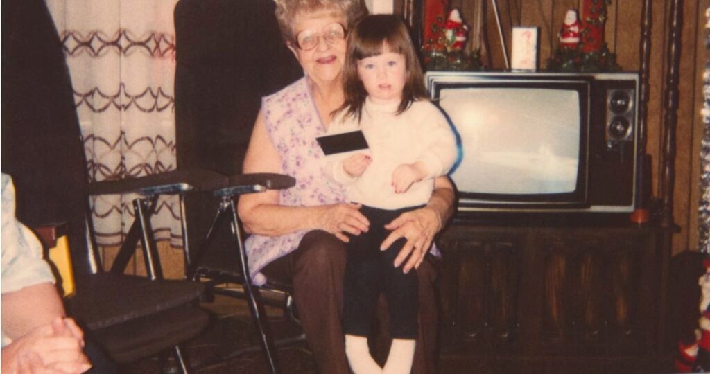 Vintage family photo of a smiling grandmother holding her young granddaughter on her lap beside an old television in a cozy, wood-paneled living room.