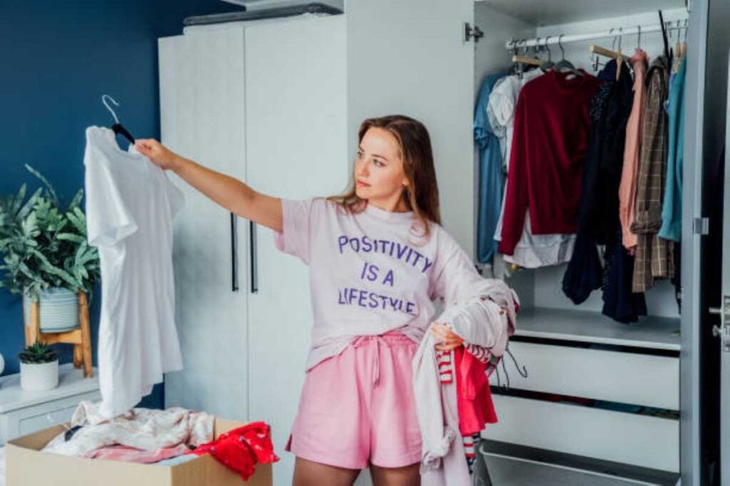 Woman decluttering her wardrobe, holding up a white shirt while carrying a pile of clothes beside an open closet.