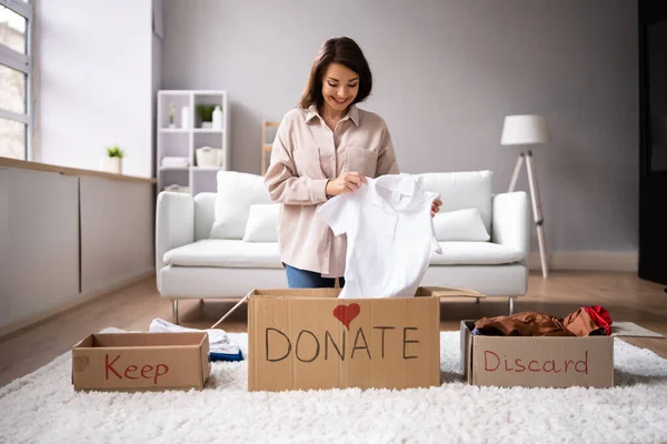 Woman sorting clothes into boxes labeled “Keep,” “Donate,” and “Discard” in a living room.