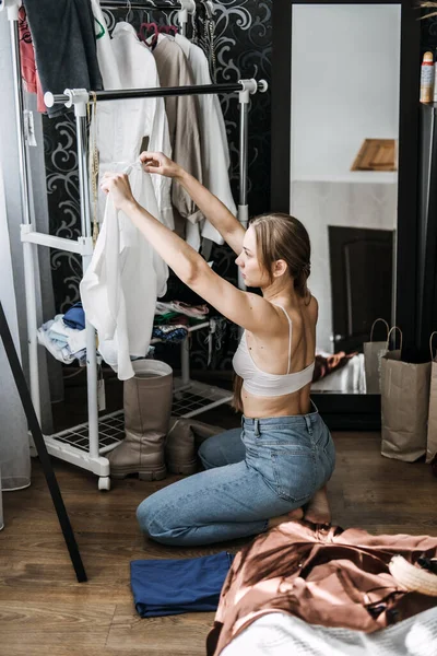 Woman kneeling beside a clothing rack while organizing white garments, with clothes scattered on the floor.