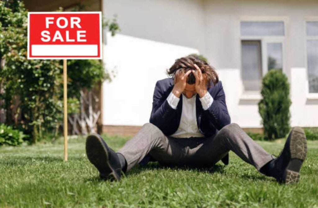 A frustrated man sits on the lawn with his head in his hands beside a “For Sale” sign in front of a house.