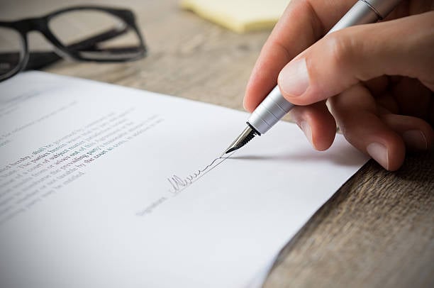 Close-up of a hand using a pen to sign a printed document on a wooden table.