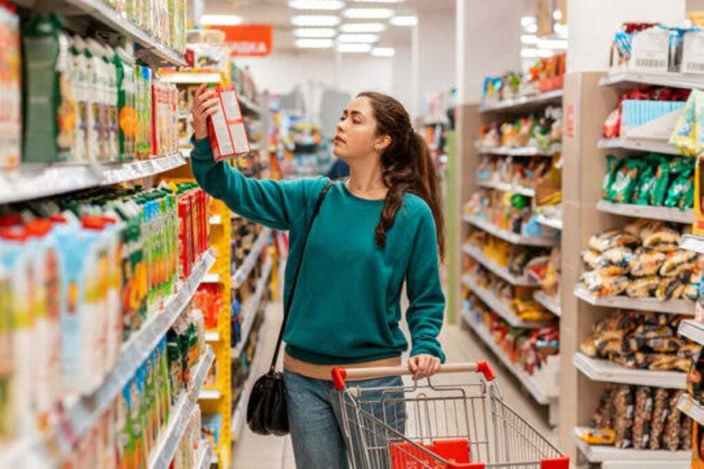 A woman shopping in a grocery store aisle examines a packaged food item while pushing a shopping cart.