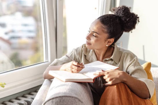 Woman sitting beside a bright window, holding a pen and looking away from an open notebook.
