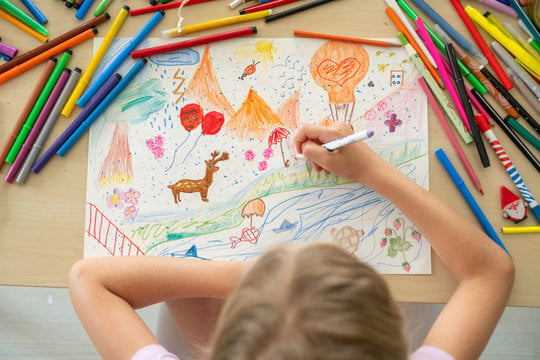Child drawing a colorful picture on paper surrounded by an assortment of colored pencils and markers.