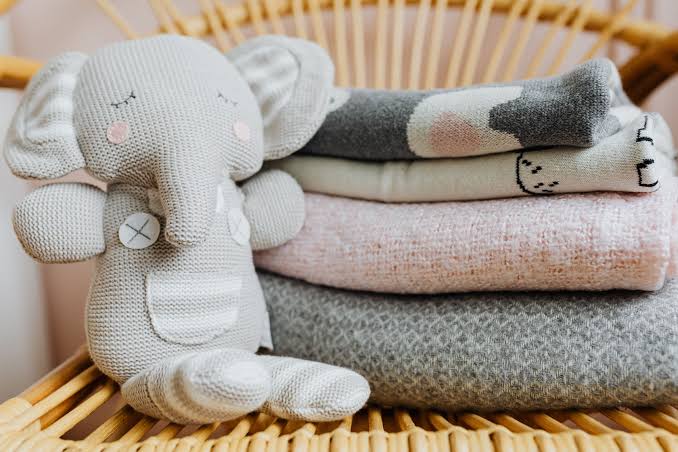 Gray knitted stuffed elephant leaning against a stack of folded blankets on a wicker chair.