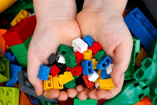 Child’s hands holding a pile of colorful plastic building bricks over a bin filled with larger blocks.