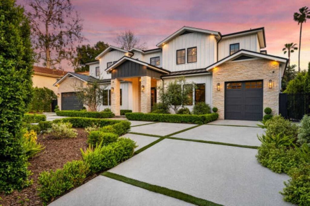 Modern two-story white house with stone accents, dark garage doors, manicured landscaping, and a wide concrete driveway at sunset.