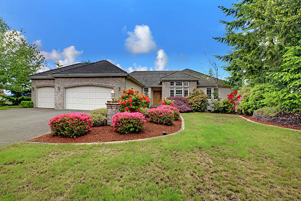 Single-story suburban brick home with a three-car garage, green lawn, mature trees, and colorful flowering shrubs in the front yard.
