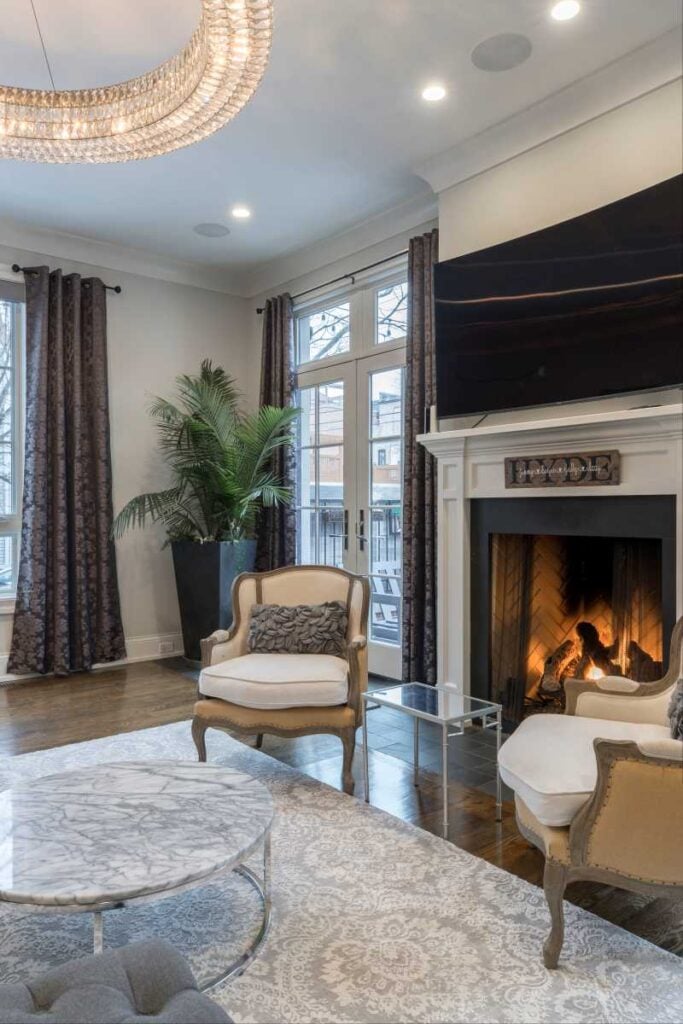 Elegant living room with a lit fireplace, large wall-mounted TV, upholstered armchairs, marble coffee table, tall windows, and a crystal chandelier.