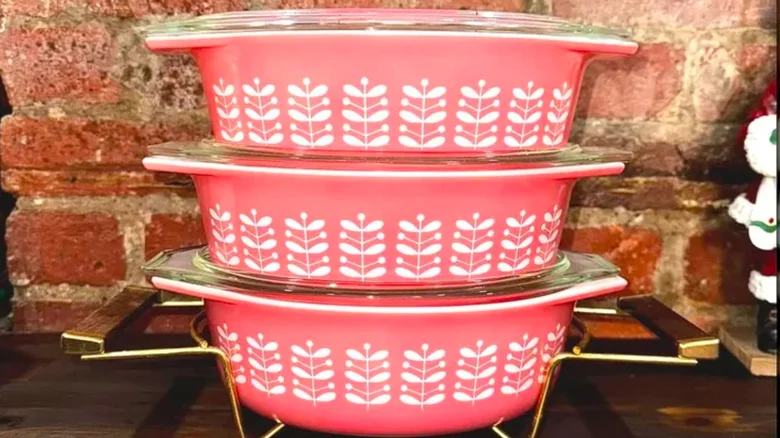 Three stacked vintage pink Pyrex casserole dishes with white leaf-and-stem patterns and clear glass lids, displayed against a brick wall.